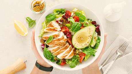 Hand holding out a bowl of salad with sliced chicken, half an avocado, and other colourful greens - Kaizen Naturals blogs