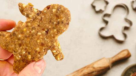 Hand holding a healthy gingerbread man cookie with baking supplies on the table in the background - Kaizen Naturals